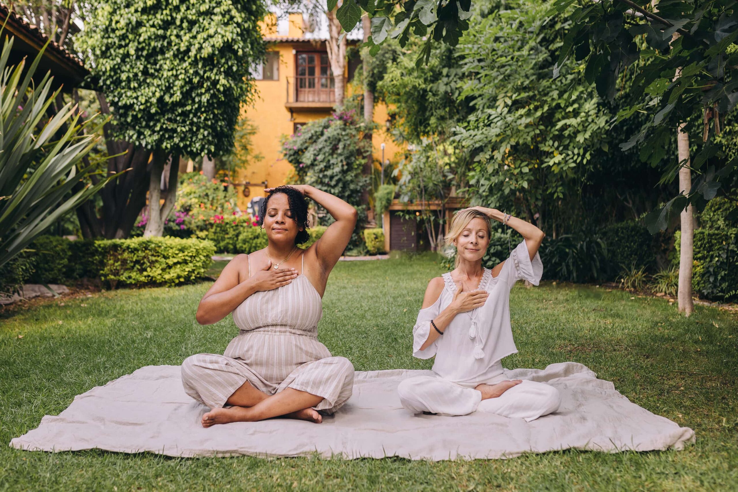 Pranayama - Unexpected Benefits of Breathing as a Spiritual Practice- Two women practicing pranayama and yoga on a mat in a garden, sitting cross-legged and performing a chest-opening pose.