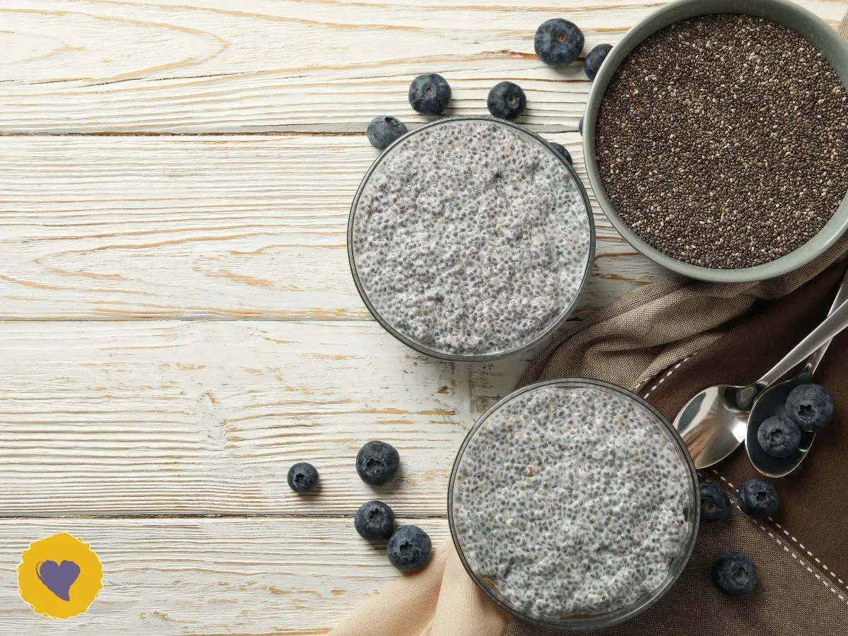 Two glasses of chia pudding topped with blueberries sit on a wooden table next to a bowl of chia seeds and two spoons. Photo from Canva Pro.