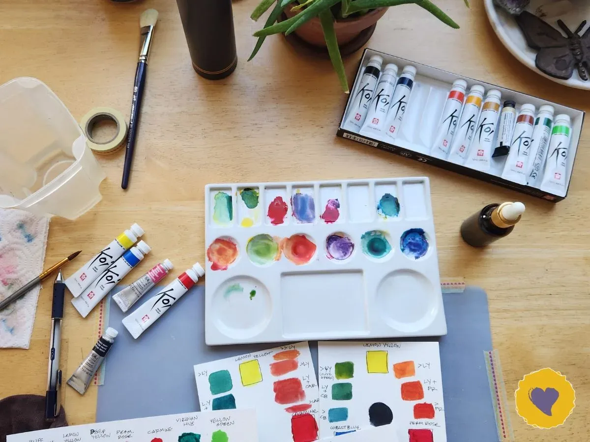 A wooden table with watercolor paints, a white palette filled with various colors, paint tubes, brushes, a plant, and color swatch papers. Photo taken by Dawn Browning