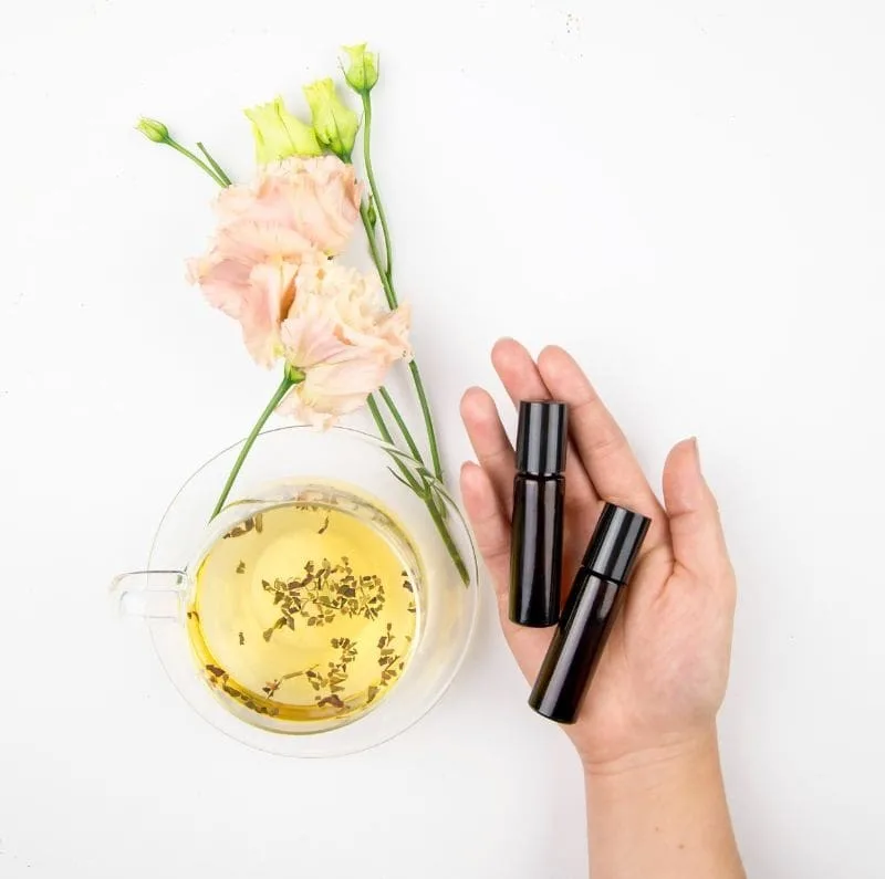 Hand holding two black bottles near a glass cup of tea with herbs, and pink flowers on a white background.